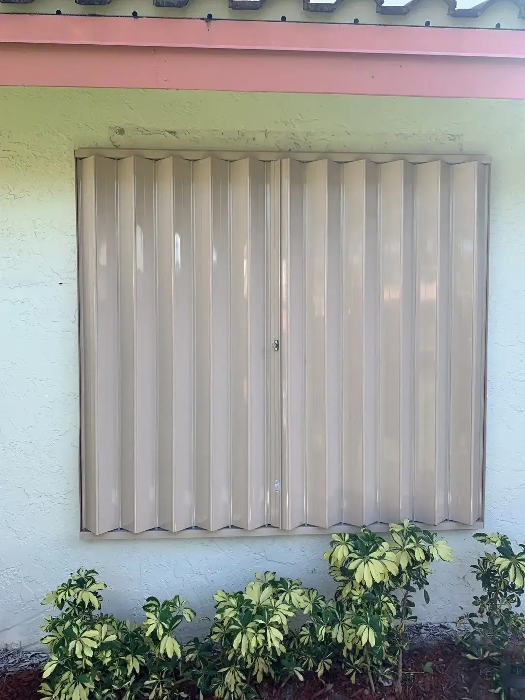 Cream accordion shutters closed over a window on a stucco South Florida wall