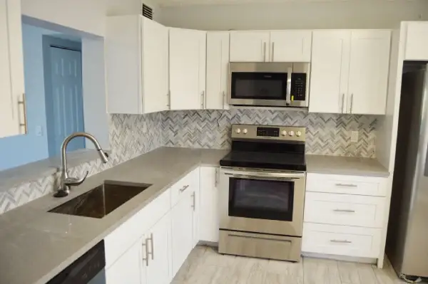 L-shaped white kitchen with gray quartz counters, mosaic backsplash and stainless appliances